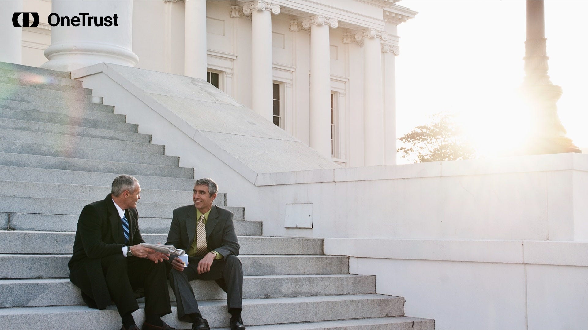 Two men in suits sit on the steps outside a neoclassical government building at sunset.