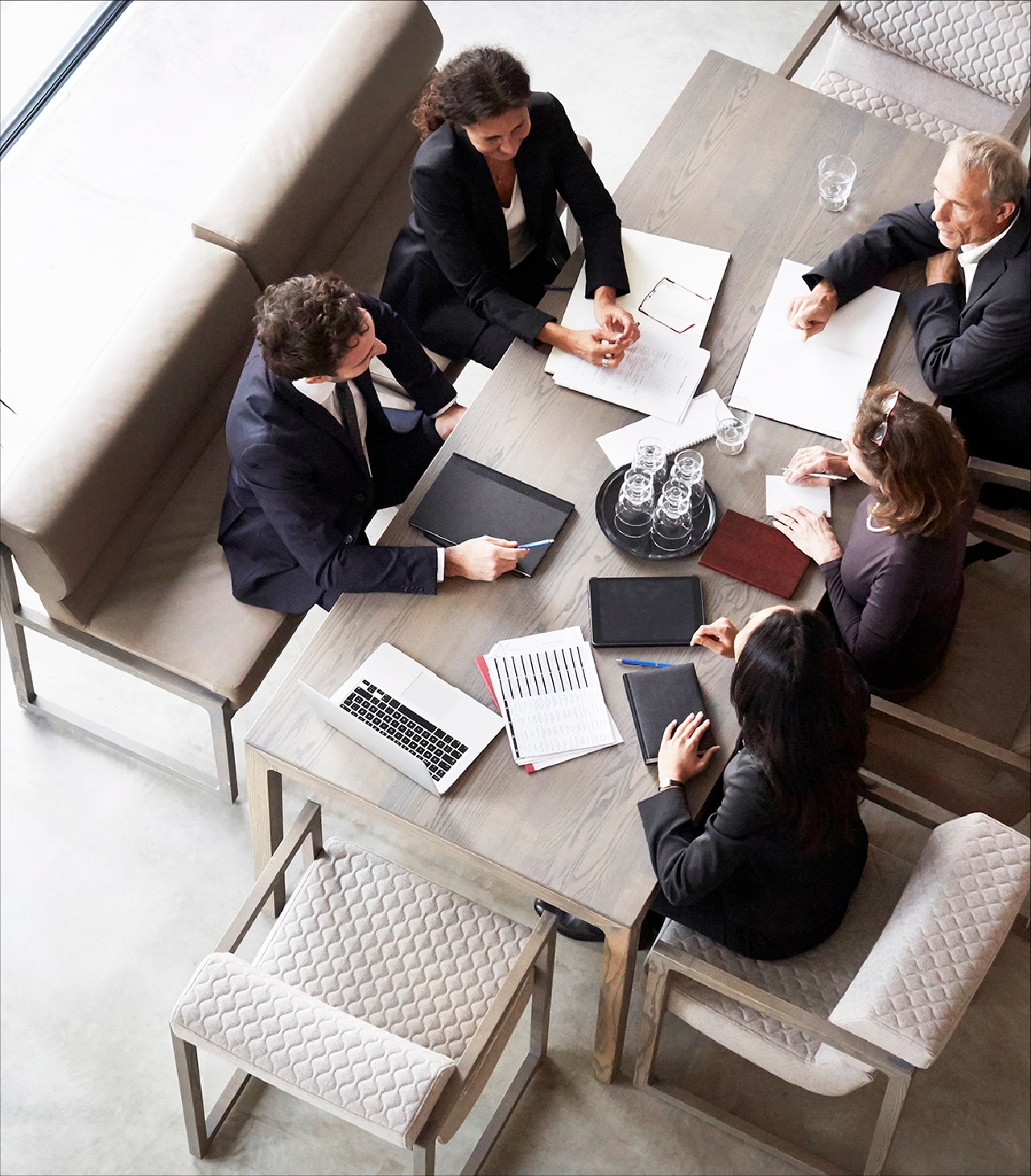 Five professionals meet around a table reviewing documents, laptops, and tablets in a modern office space.